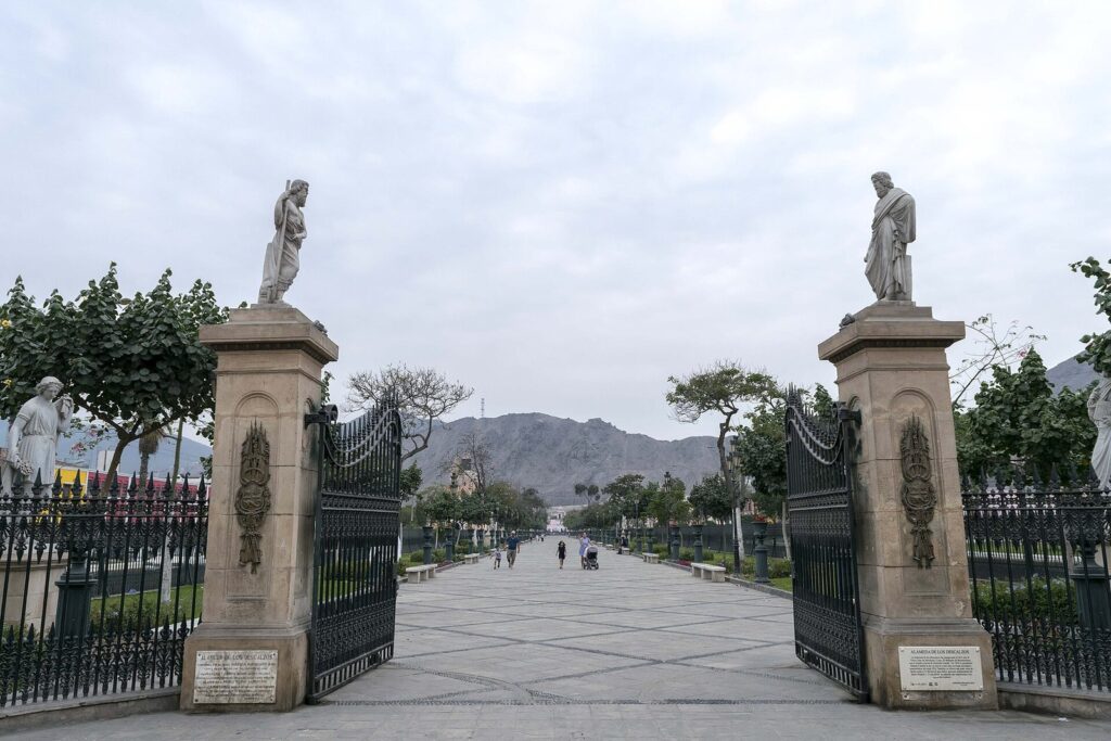 Stone gateway with marble statues at the entrance to the Alameda de los Descalzos promenade in Rímac, Lima, with dry hills behind