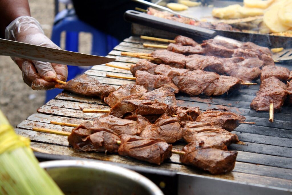 Skewers of anticuchos, grilled beef heart, cooking over a street-food grill in Lima, Peru, as a vendor slices the meat