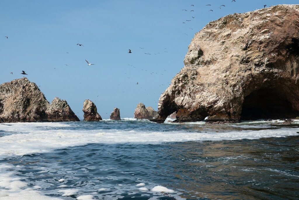 Guano-streaked rock arches and sea stacks of the Ballestas Islands off Paracas, Peru, with seabirds wheeling over a blue sea