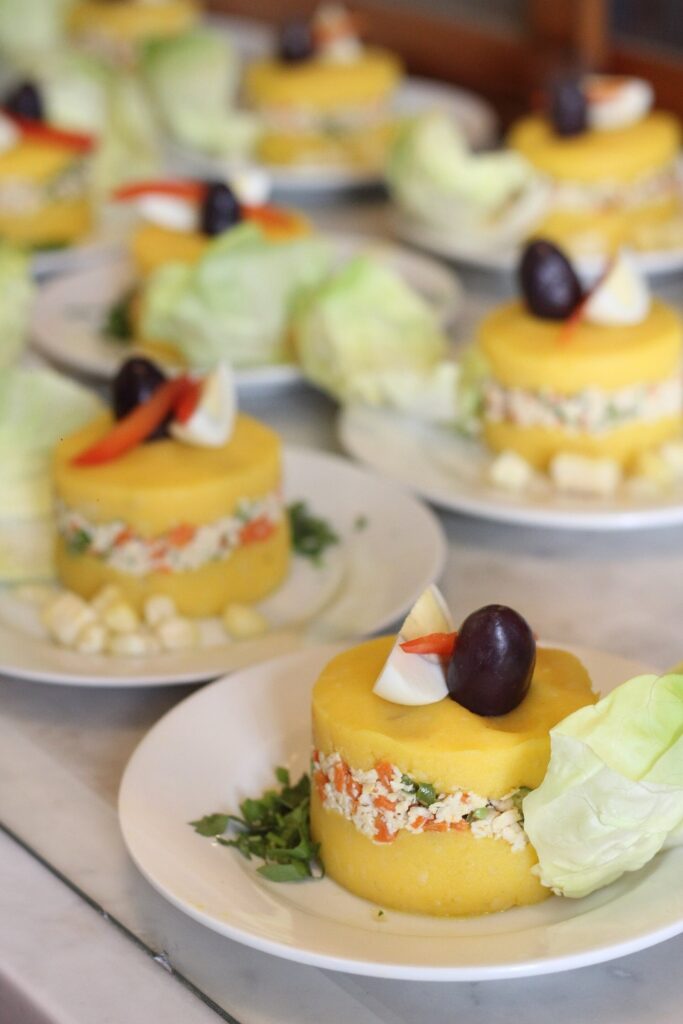 Rows of individual causa rellena portions, yellow potato layers with a chicken and vegetable filling, topped with black olives and slices of egg