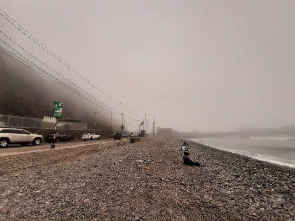Low fog over the pebble beach at Playa Makaha below the Miraflores cliffs, with the coastal road fading into mist
