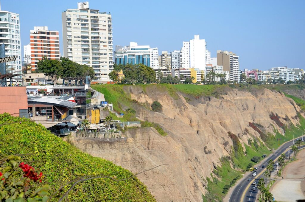 Larcomar shopping center built into the cliff at Miraflores, Lima, with high-rise towers above and the Costa Verde road below under a blue sky