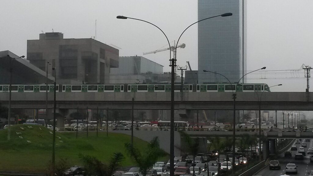 A Metro Line 1 train crossing its elevated viaduct above a busy avenue with traffic and office buildings in Lima