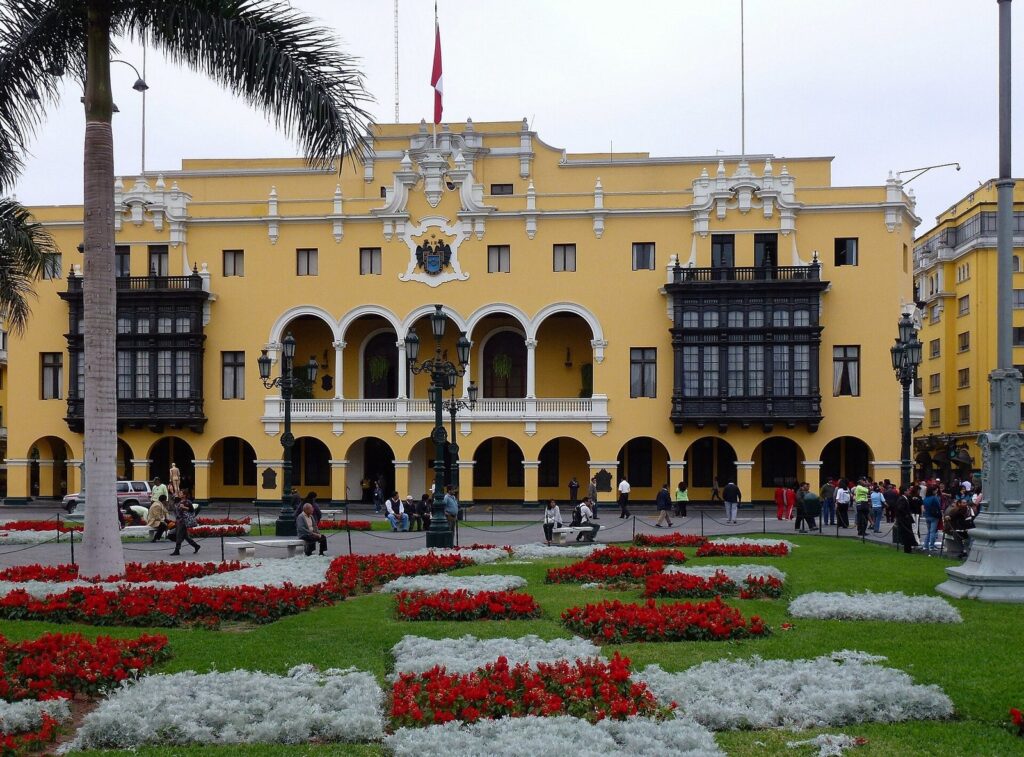 Yellow Municipal Palace with dark carved wooden balconies behind red flower beds and palm trees on the Plaza Mayor of Lima