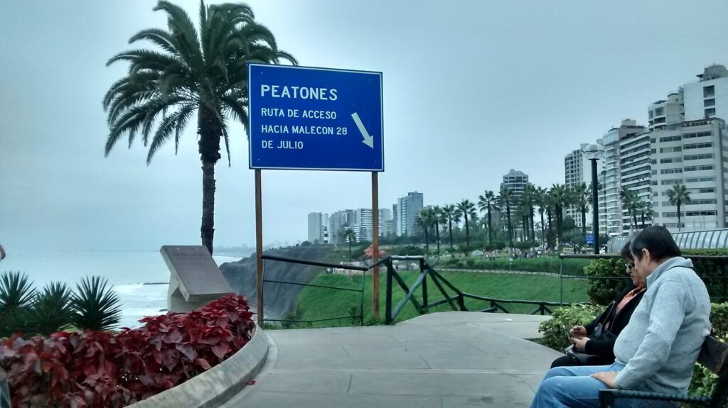 Palm-lined clifftop walkway on the Malecón de Miraflores in Lima with a blue pedestrian sign and apartment towers along the coast
