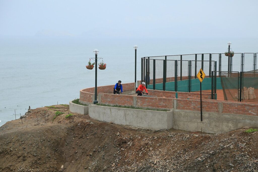 Two cyclists on a clifftop bike path with a brick wall and sports courts above the sea in Miraflores, Lima