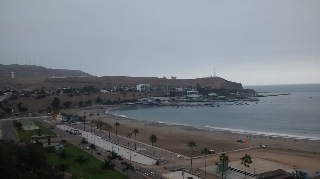 Wide view of the sandy Playa Agua Dulce in Chorrillos, Lima, with a palm-lined promenade, fishing boats in the bay and the Morro Solar headland
