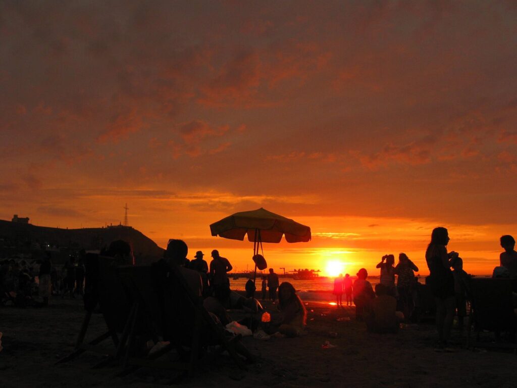 Orange sunset over Playa Agua Dulce in Chorrillos, Lima, with beachgoers and a sun umbrella in silhouette