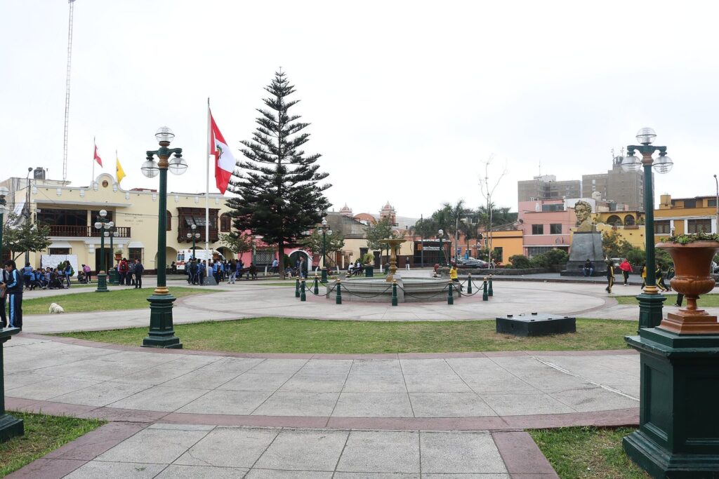 Plaza Bolívar in Pueblo Libre, Lima, with lamp posts, a tall pine, flags and low colonial-style buildings around the square
