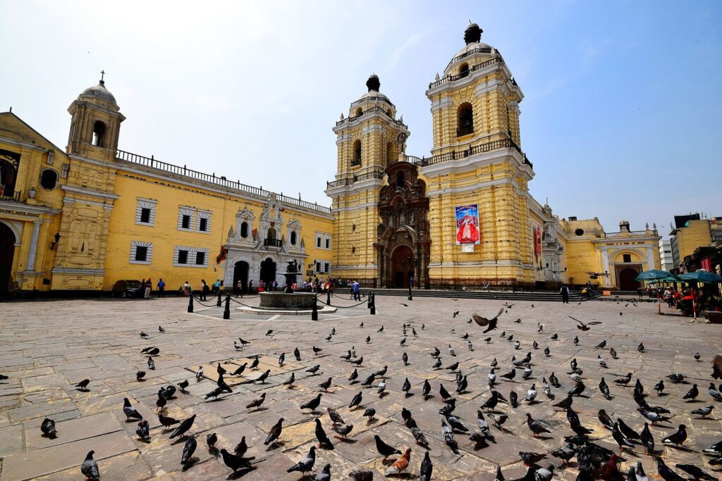 Yellow baroque facade and twin towers of the Basilica and Convent of San Francisco in Lima's historic center, with pigeons on the plaza under a blue sky